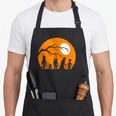 Trick halloween apron - Grey silhouettes holding pumpkin buckets inside an orange circle with a white moon and tree — shown on a black cotton canvas apron against a white background