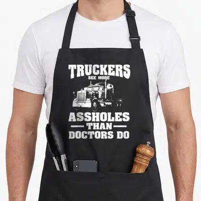 See semi truck apron - White semi truck graphic with stacked text TRUCKERS SEE MORE ASSHOLES THAN DOCTORS DO — shown on a black cotton canvas apron against a white background