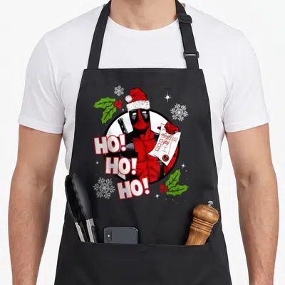 List santa apron - Deadpool in Santa hat holding Naughty List YOU with HO HO HO text and snowflakes — shown on a black cotton canvas apron against a white background