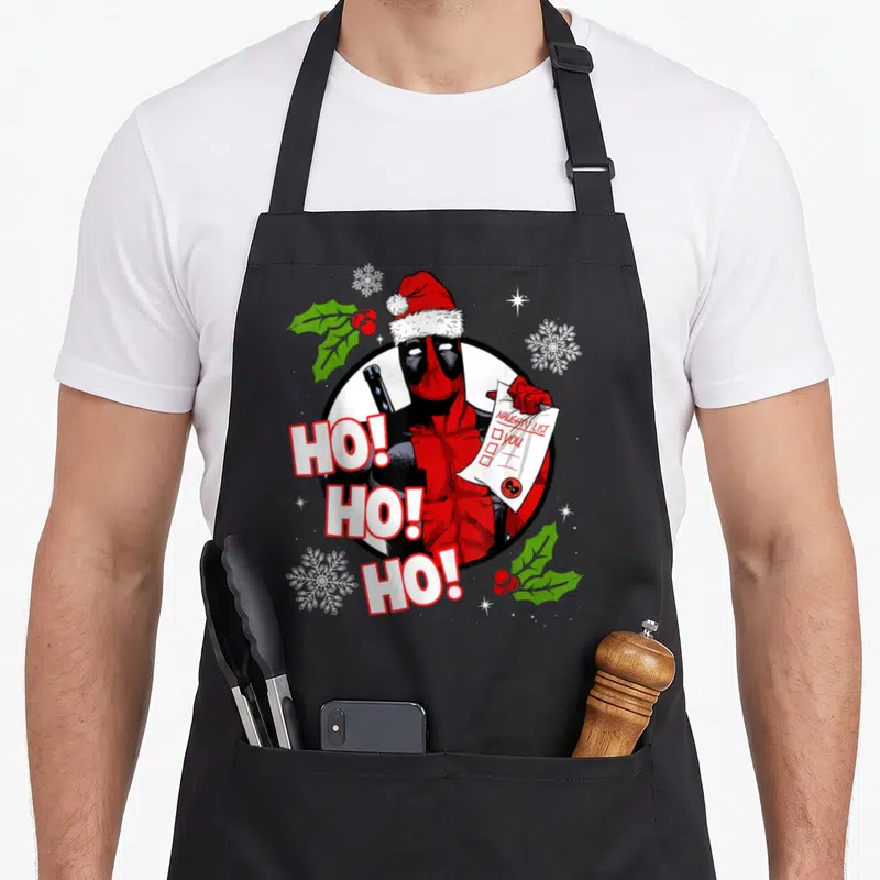 List santa apron - Deadpool in Santa hat holding Naughty List YOU with HO HO HO text and snowflakes — shown on a black cotton canvas apron against a white background
