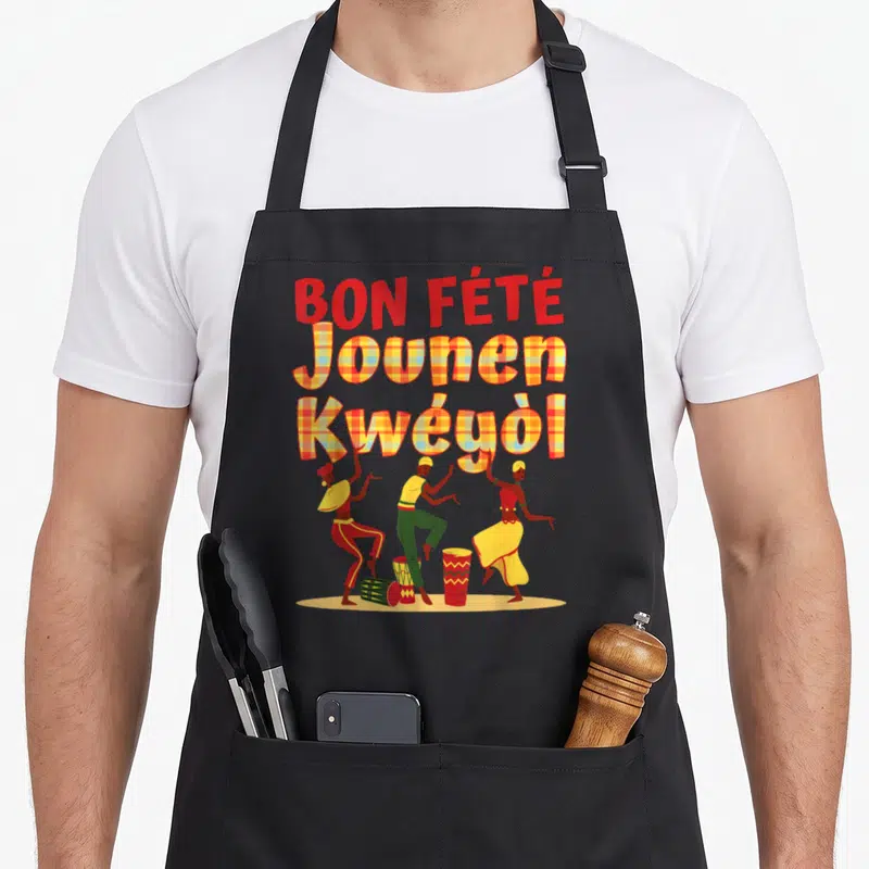 Bon the plaid apron - BON FETE Jounen Kweyol text in plaid above three dancers and drums on yellow ground — shown on a black cotton canvas apron against a white background