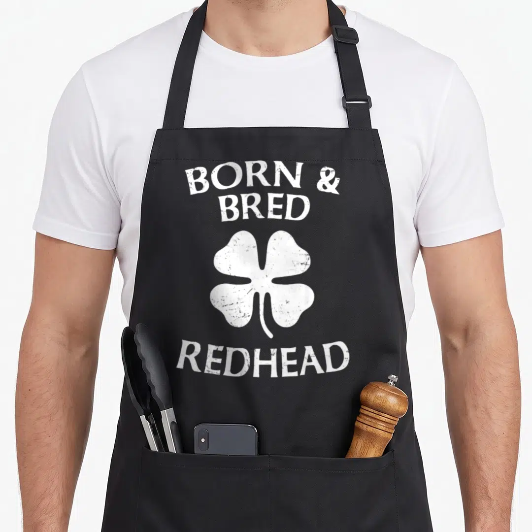 Bred st patrick day aprons - White distressed four-leaf clover between Born & Bred and Redhead text — shown on a black cotton canvas apron against a white background