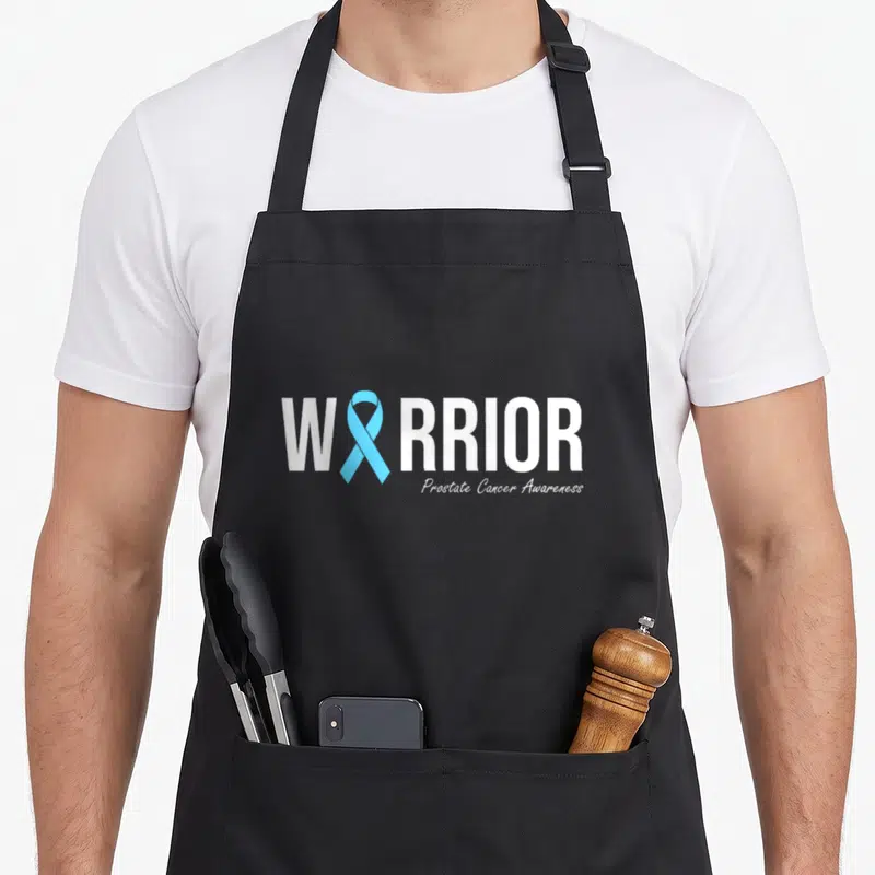 Prostate family aprons - WARRIOR text with light blue ribbon replacing letter A and Prostate Cancer Awareness below — shown on a black cotton canvas apron against a white background