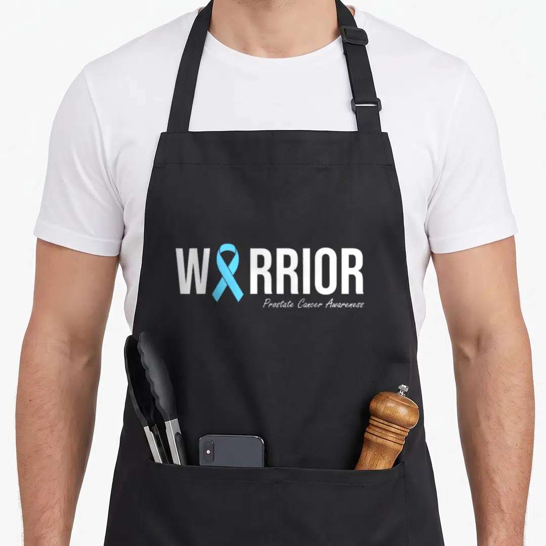 Prostate family aprons - WARRIOR text with light blue ribbon replacing letter A and Prostate Cancer Awareness below — shown on a black cotton canvas apron against a white background