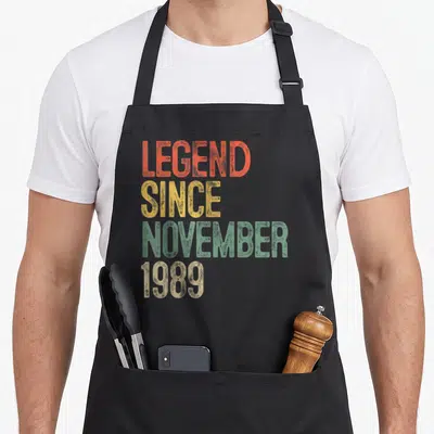 November 30th birthday apron - Distressed stacked text LEGEND SINCE NOVEMBER 1989 in orange yellow green beige — shown on a black cotton canvas apron against a white background