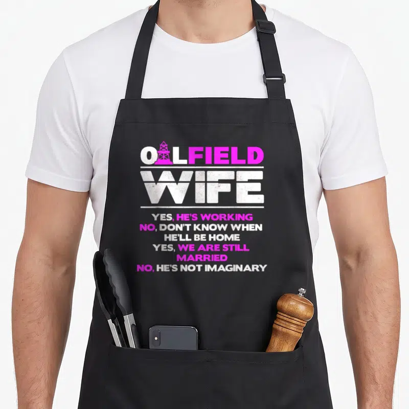 Roughneck wife apron - Pink and white OILFIELD WIFE text with oil rig inside O and stacked yes no answers — shown on a black cotton canvas apron against a white background