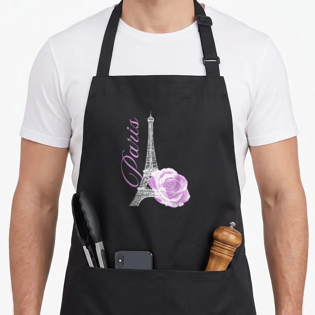Souvenir paris apron - White Eiffel Tower with vertical pink Paris text and overlapping pink rose — shown on a black cotton canvas apron against a white background