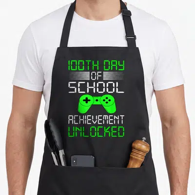 School video game apron - Green game controller between 100th day of school and achievement unlocked text — shown on a black cotton canvas apron against a white background
