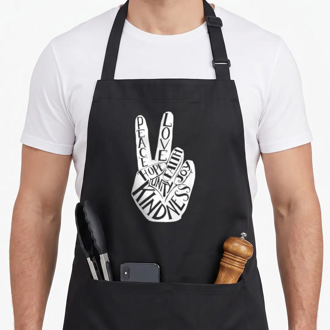 Anti unity day apron - White hand peace sign formed by words PEACE LOVE UNITY KINDNESS — shown on a black cotton canvas apron against a white background