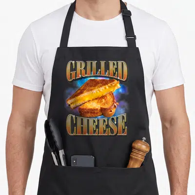 Grilled gag aprons - GRILLED CHEESE text framing a toasted sandwich over a blue space nebula background — shown on a black cotton canvas apron against a white background