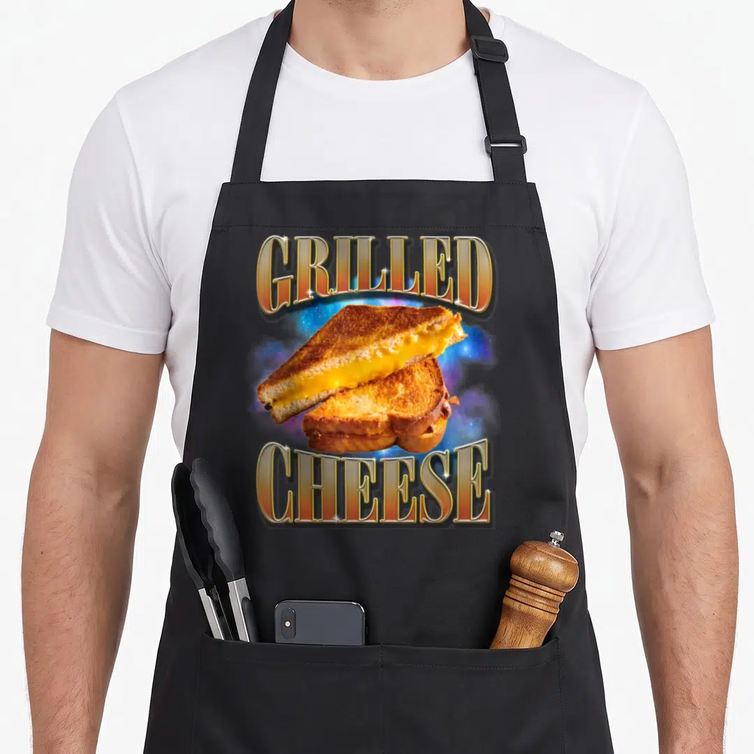 Grilled gag aprons - GRILLED CHEESE text framing a toasted sandwich over a blue space nebula background — shown on a black cotton canvas apron against a white background