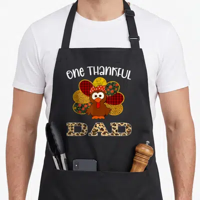 Cute thankful thanksgiving apron - Cartoon turkey with patterned feathers and one THANKFUL DAD text in leopard print — shown on a black cotton canvas apron against a white background