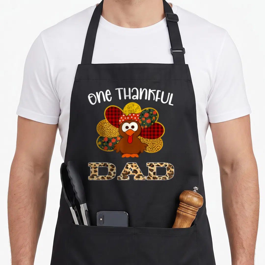 Cute thankful thanksgiving apron - Cartoon turkey with patterned feathers and one THANKFUL DAD text in leopard print — shown on a black cotton canvas apron against a white background