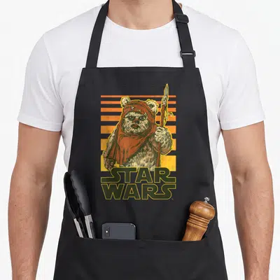 Wicket sunset apron - Ewok holding spear, STAR WARS text, orange yellow stripes, distressed print — shown on a black cotton canvas apron against a white background