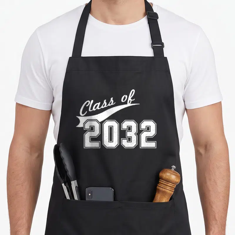 Kindergarten class funny apron - White Class of 2032 text on ribbon banner with block numerals — shown on a black cotton canvas apron against a white background