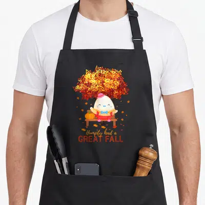 Thanksgiving autumn cute fall aprons - Cartoon egg character on bench under autumn tree with pumpkin and text Humpty — shown on a black cotton canvas apron against a white background