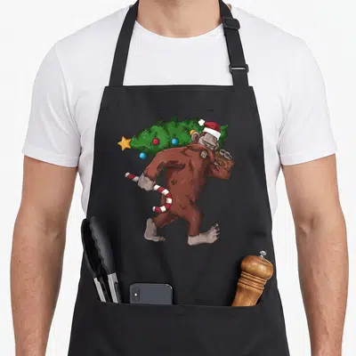 Bigfoot womens christmas aprons - Walking brown bigfoot carrying green tree and red white candy cane with Santa hat — shown on a black cotton canvas apron against a white background