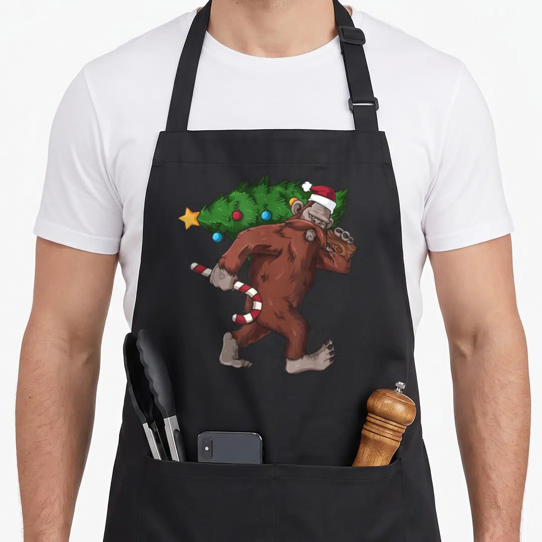 Bigfoot womens christmas aprons - Walking brown bigfoot carrying green tree and red white candy cane with Santa hat — shown on a black cotton canvas apron against a white background