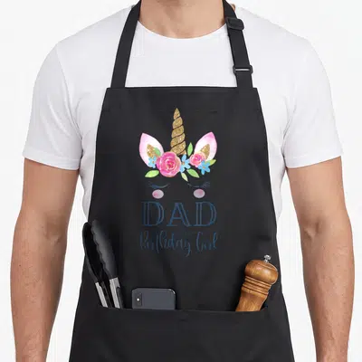 Funny birthday unicorn apron - Unicorn head with gold horn, pink flowers, and blue DAD of the Birthday Girl text — shown on a black cotton canvas apron against a white background