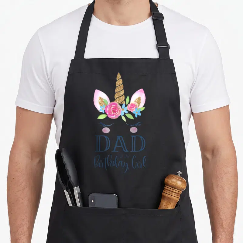 Funny birthday unicorn apron - Unicorn head with gold horn, pink flowers, and blue DAD of the Birthday Girl text — shown on a black cotton canvas apron against a white background