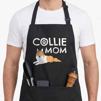 Collie puppy dog apron - Cartoon collie lying below white text COLLIE MOM with paw print in O — shown on a black cotton canvas apron against a white background