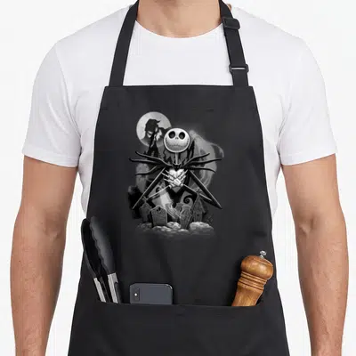 Night christmas apron - Jack Skellington skeleton standing in a graveyard with tombstones under a full moon in black — shown on a black cotton canvas apron against a white background