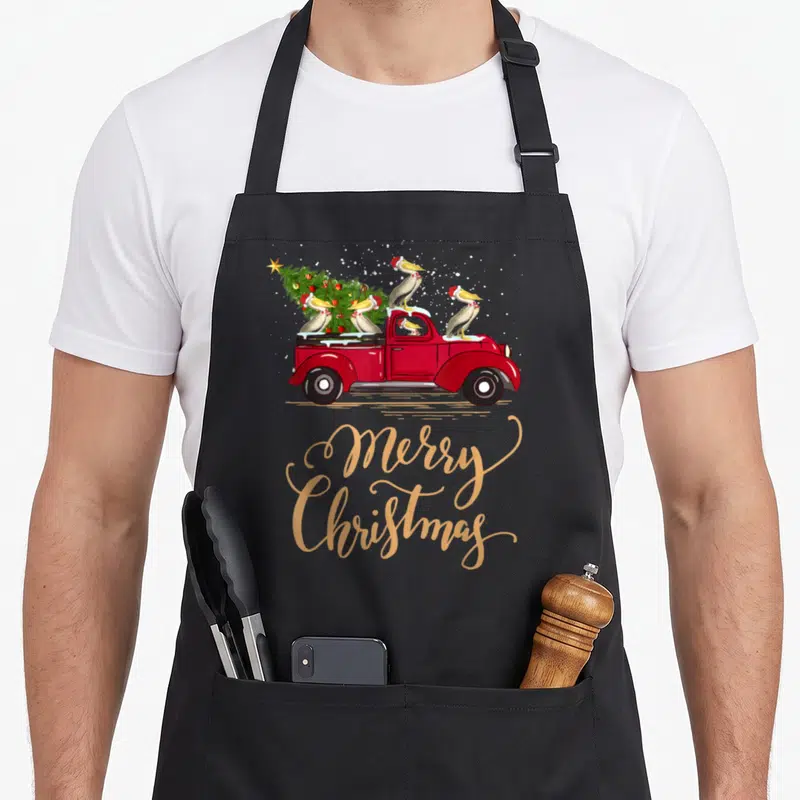 Drive christmas tree apron - Red truck carrying Christmas tree and five pelicans with gold Merry Christmas script — shown on a black cotton canvas apron against a white background