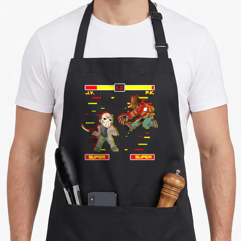 Horror funny halloween aprons - Two horror characters facing off with K.O and SUPER text in red yellow and green — shown on a black cotton canvas apron against a white background