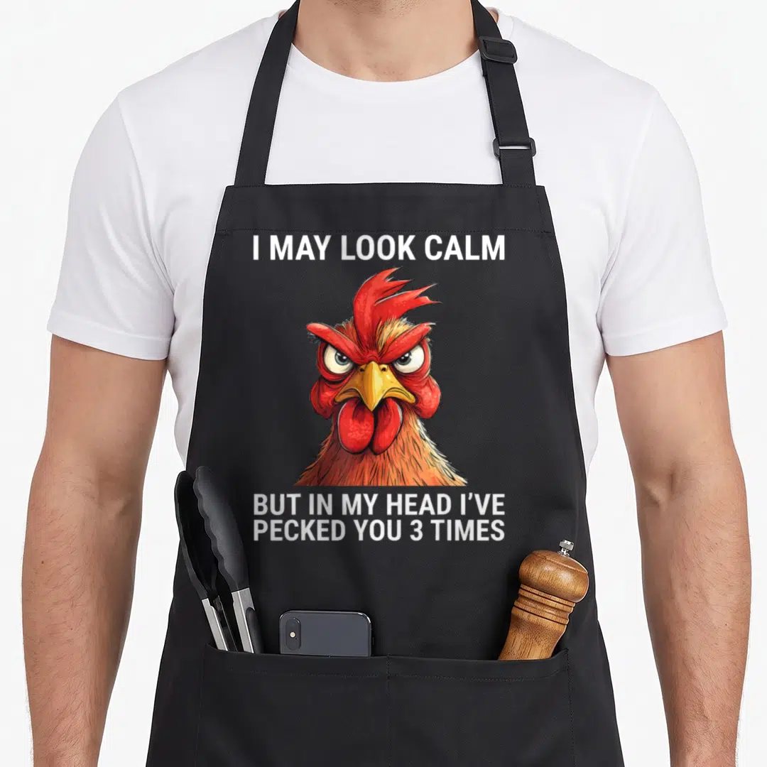 May but i love my rooster apron - Cartoon rooster head in red and orange with white text reading I may look calm — shown on a black cotton canvas apron against a white background