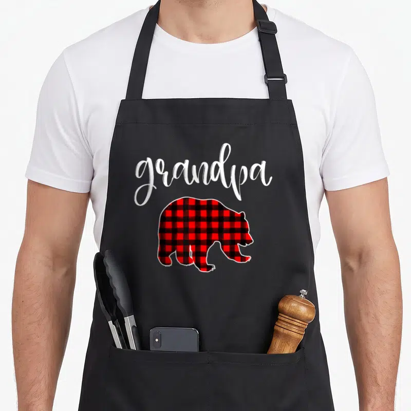Family eve buffalo plaid christmas apron - Grandpa script text above red and black plaid bear silhouette — shown on a black cotton canvas apron against a white background