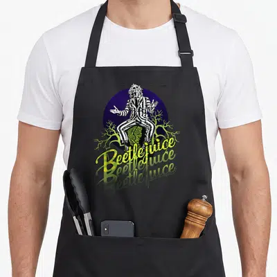 Three portrait novelty apron - Beetlejuice sitting on tombstone with purple circle and lime green branches and yellow — shown on a black cotton canvas apron against a white background