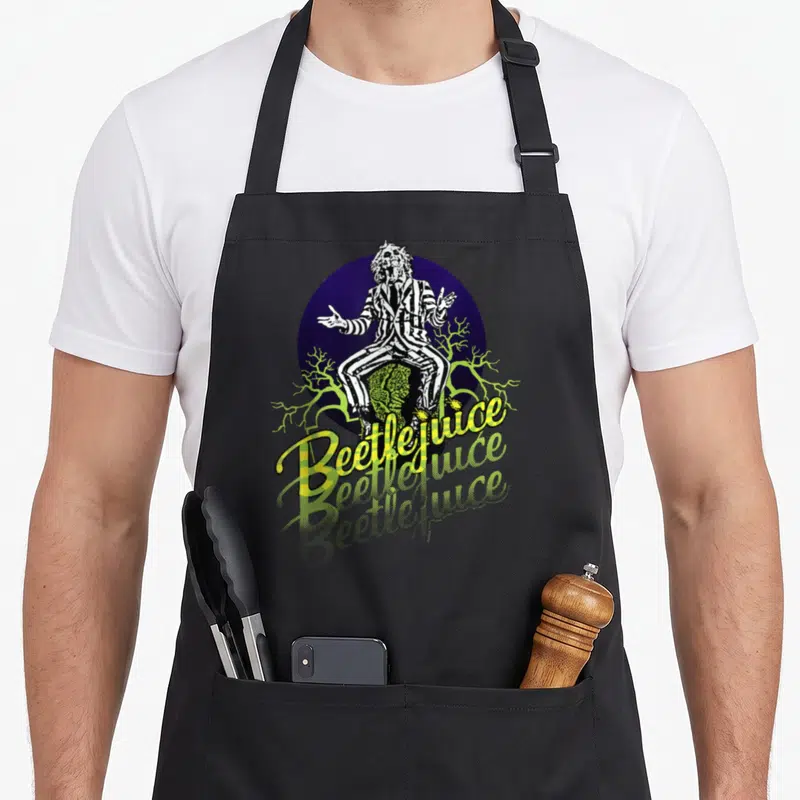 Three portrait novelty apron - Beetlejuice sitting on tombstone with purple circle and lime green branches and yellow — shown on a black cotton canvas apron against a white background