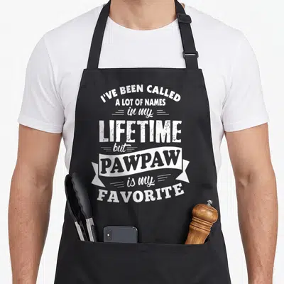 Called lot of aprons - White typography reading I've Been Called a Lot of Names with PAWPAW inside a ribbon banner — shown on a black cotton canvas apron against a white background