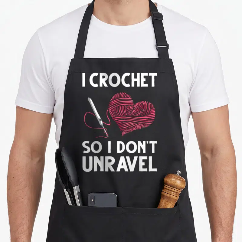 Crocheter crochet apron - White text I Crochet So I Don't Unravel around pink yarn heart and hook — shown on a black cotton canvas apron against a white background