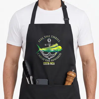 Feel fishing aprons - Realistic mahi mahi in yellow and green over white anchor with arched text FEEL THE THRILL CATCH — shown on a black cotton canvas apron against a white background