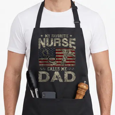 Flag nurses apron - Distressed beige text reading My Favorite Nurse Calls Me Dad over a USA flag with a stethoscope — shown on a black cotton canvas apron against a white background