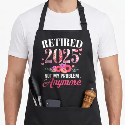 Problem floral apron - Retired 2025 text with pink floral fill and hearts, not my problem anymore script — shown on a black cotton canvas apron against a white background