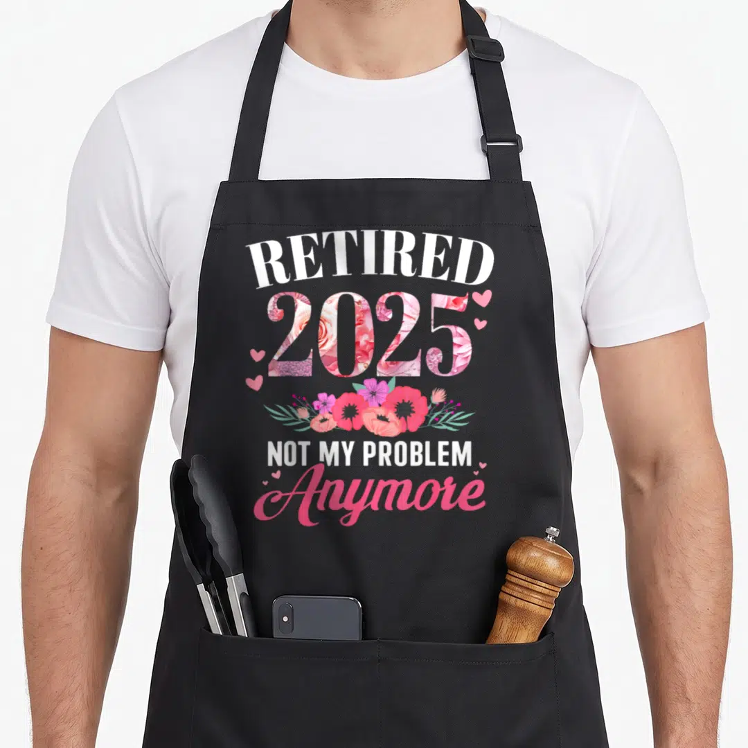 Problem floral apron - Retired 2025 text with pink floral fill and hearts, not my problem anymore script — shown on a black cotton canvas apron against a white background