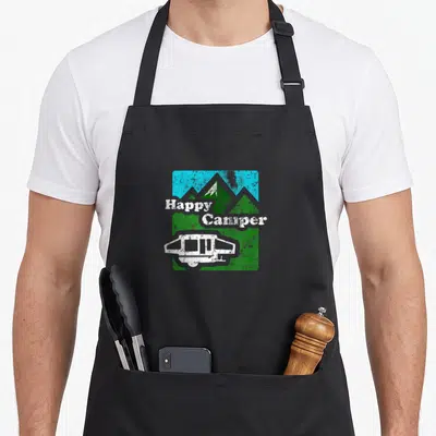 Popup camping apron - Happy Camper text over green mountains and blue sky with a white pop-up trailer in a distressed — shown on a black cotton canvas apron against a white background