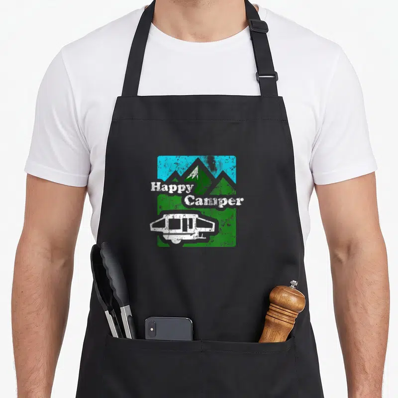 Popup camping apron - Happy Camper text over green mountains and blue sky with a white pop-up trailer in a distressed — shown on a black cotton canvas apron against a white background