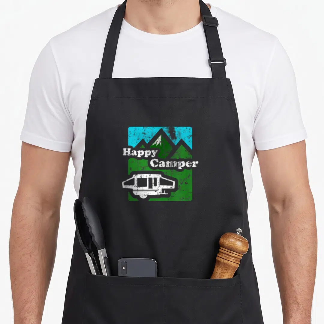 Popup camping apron - Happy Camper text over green mountains and blue sky with a white pop-up trailer in a distressed — shown on a black cotton canvas apron against a white background