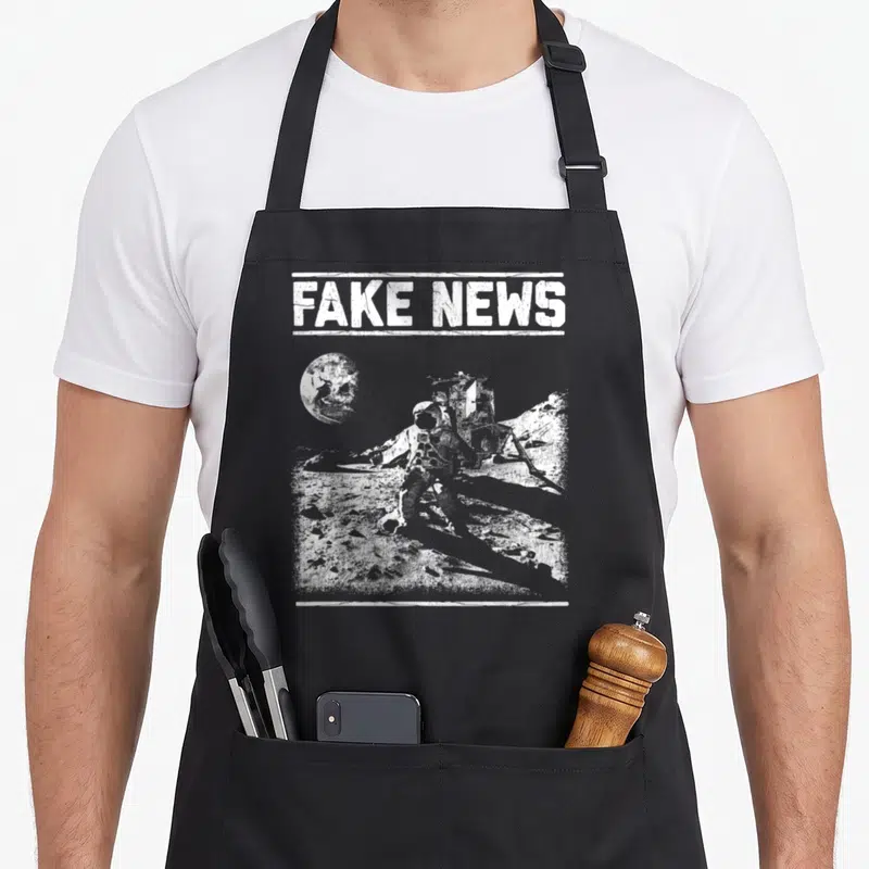 Moon apollo apron - FAKE NEWS text above distressed white astronaut and lunar module on moon surface — shown on a black cotton canvas apron against a white background