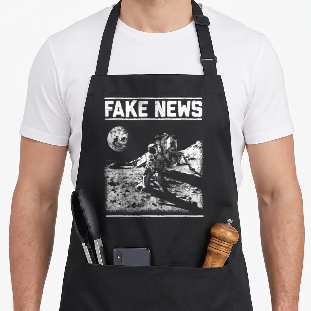 Moon apollo apron - FAKE NEWS text above distressed white astronaut and lunar module on moon surface — shown on a black cotton canvas apron against a white background