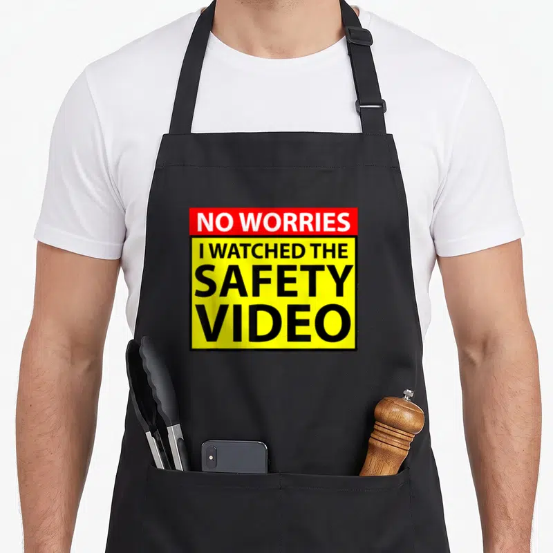 Worry safety apron - NO WORRIES I WATCHED THE SAFETY VIDEO text in red and yellow rectangles — shown on a black cotton canvas apron against a white background