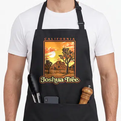 Wpa tree apron - Retro California Joshua Tree landscape with orange sky, brown mountains, and stylized trees — shown on a black cotton canvas apron against a white background