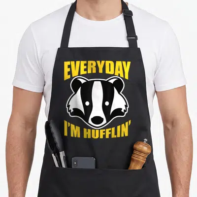 Everyday badger apron - Yellow text reading Everyday Im Hufflin around black and white badger head — shown on a black cotton canvas apron against a white background