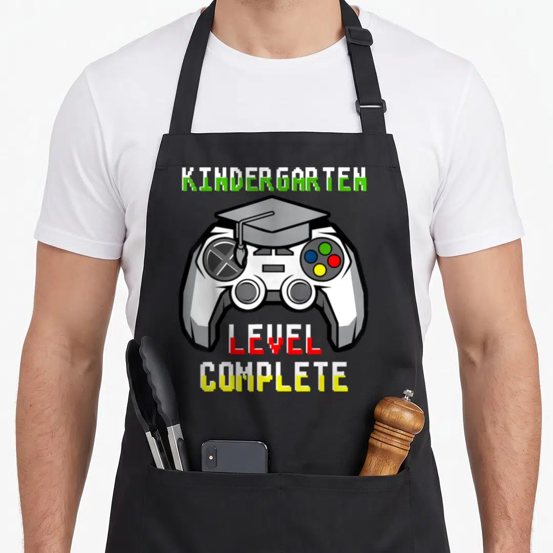 Kindergarten video game apron - Controller with graduation cap and pixelated text reading KINDERGARTEN LEVEL COMPLETE — shown on a black cotton canvas apron against a white background