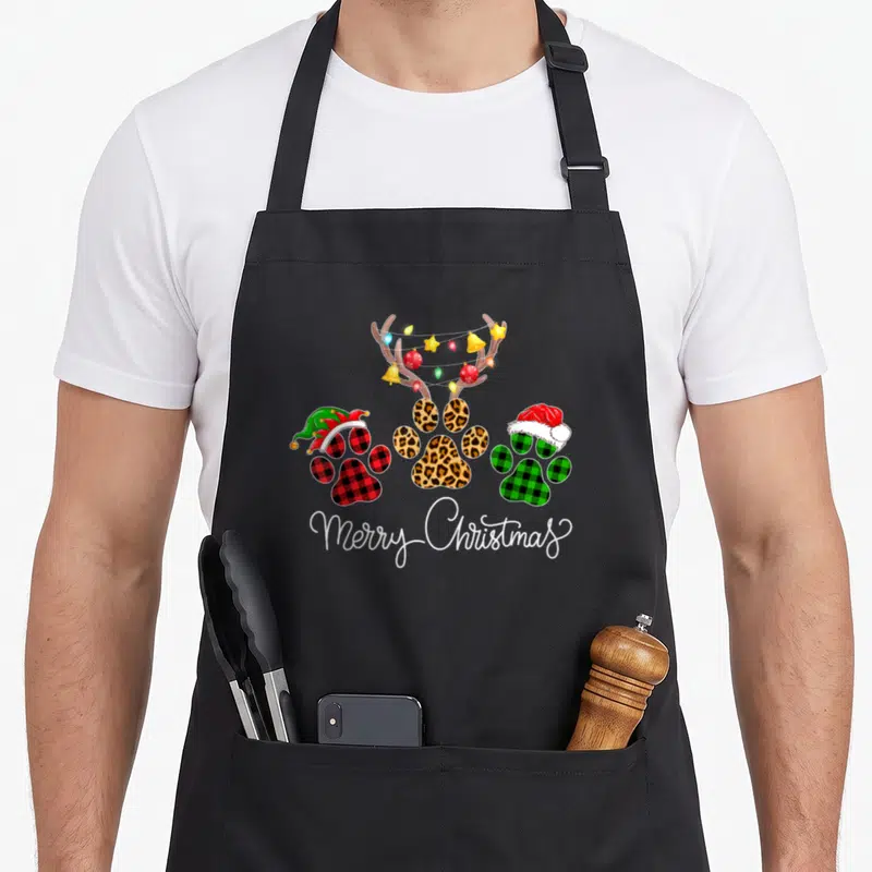 Dog buffalo plaid christmas apron - Three paw prints in plaid and leopard with antlers, lights, hats, and Merry — shown on a black cotton canvas apron against a white background