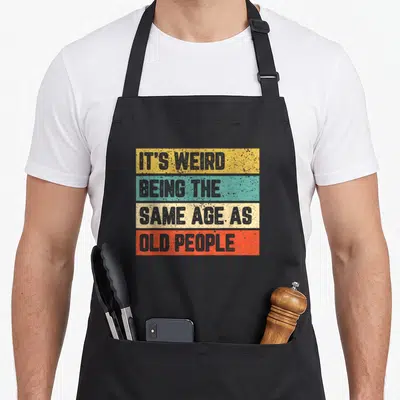 Cool general weird aprons - Stacked yellow teal beige and orange bars with grey text reading Its Weird Being The Same — shown on a black cotton canvas apron against a white background