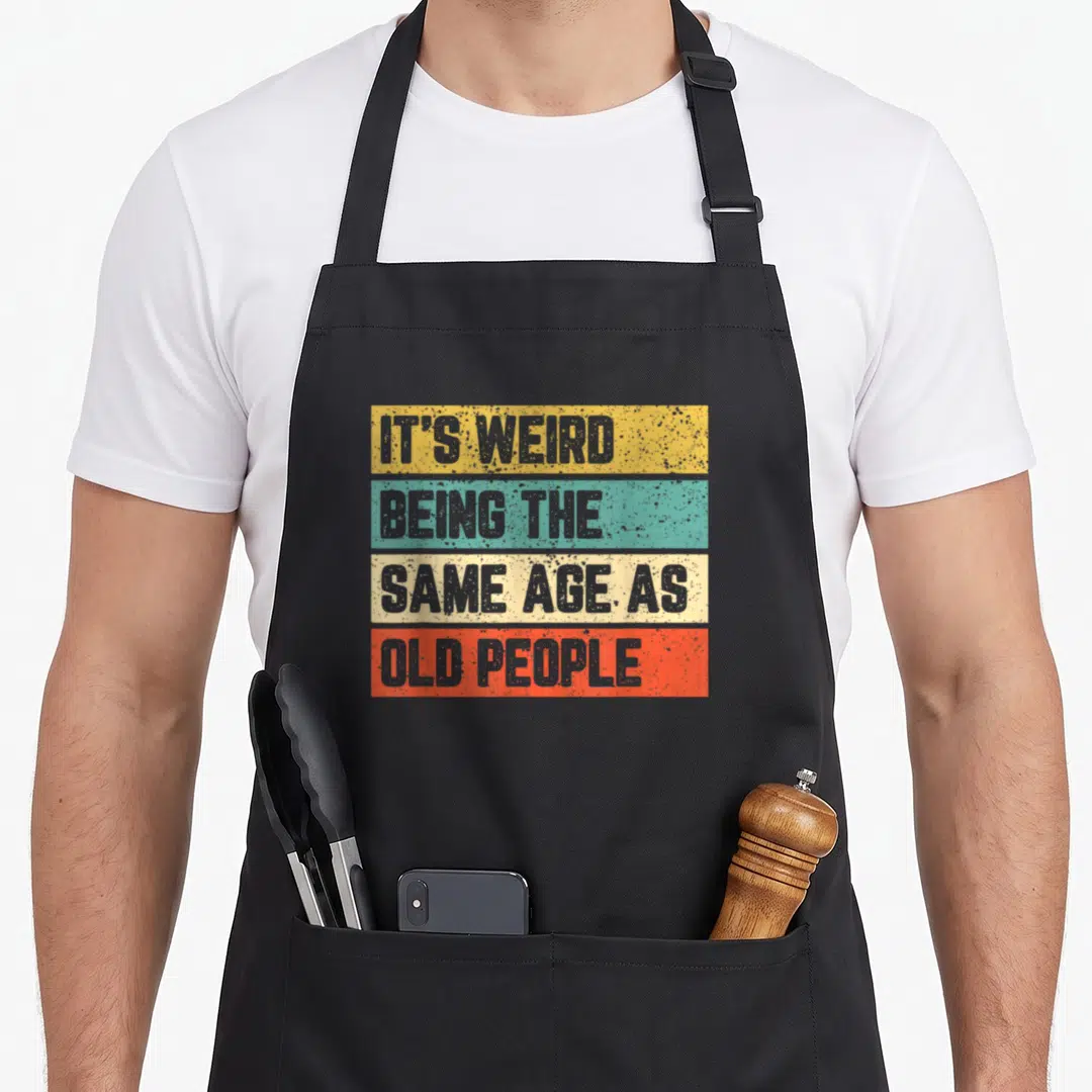 Cool general weird aprons - Stacked yellow teal beige and orange bars with grey text reading Its Weird Being The Same — shown on a black cotton canvas apron against a white background